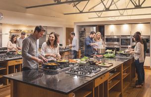 Wide shot of the open-plan Dublin Cookery School kitche, Couples work at individual stations with steam rising from pans on gas hobs, while tutors circulate to provide hands-on guidance.