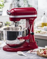 A red KitchenAid Artisan stand mixer in a professional kitchen setting, with a dough hook inside a stainless steel bowl and a fresh pizza being prepared in the background.