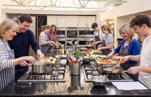 A group of mothers cook with their children collaboratively around a central kitchen island unit. Participants prepare food and stir pans on hobs in a bright, naturally lit space.
