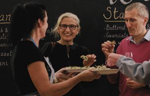 A group of attendees standing and socialising while eating appetisers from small plates during a cookery event, with a handwritten menu on a chalkboard in the background.
