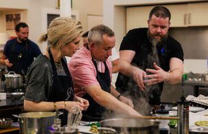 A group of students in black aprons collaborating over a steaming pot during a hands-on cookery class, listening to instruction in a busy kitchen environment.