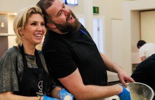 Close-up of two friends laughing heartily while mixing ingredients in a stainless steel bowl. The image captures the social and enjoyable atmosphere of a practical cookery class.