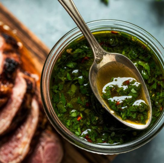 Close-up overhead shot of fresh chimichurri sauce in a glass jar with a vintage silver spoon, served alongside sliced grilled meat on a wooden board