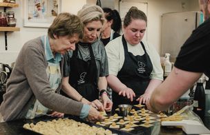 Three students wearing black Dublin Cookery School aprons stand at a granite worktop, shaping fresh pasta into tortellini under the supervision of a tutor.