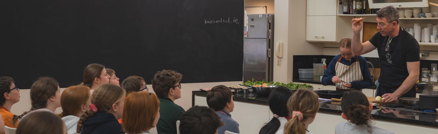 Professional tutor demonstrating a cooking technique to a group of young students in the demo kitchen during a Growing Gourmets summer camp at Dublin Cookery School, Blackrock