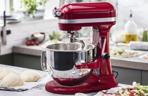 An Empire Red KitchenAid Artisan stand mixer on a grey countertop in a bright kitchen. The dough hook is attached, and three smooth, round balls of pizza dough are resting on a floured surface to the left. In the background, fresh ingredients and prepared pizza bases are visible.