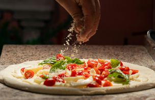 A close-up, atmospheric shot of a hand sprinkling grated parmesan cheese over a fresh pizza. The pizza is topped with halved cherry tomatoes, fresh basil leaves, and slices of mozzarella on a floured stone surface.