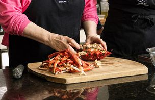 Cook in an apron preparing a crab on a wooden board at a kitchen counter.