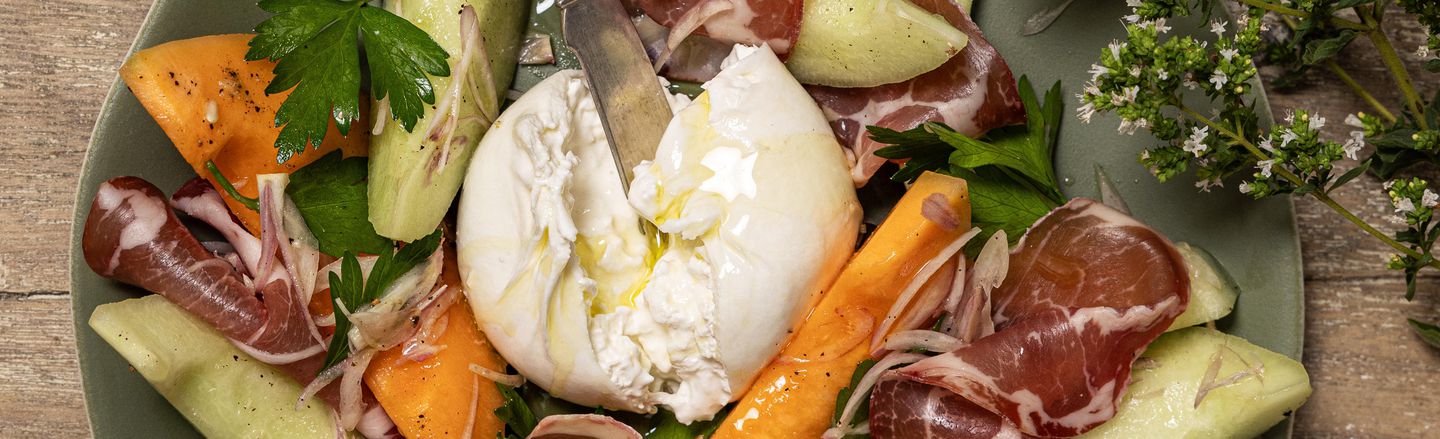 Overhead view of a salad served on a green plate, centring on a torn ball of burrata cheese drizzled with olive oil and topped with an ornate silver knife. Surrounding the cheese are wedges of cantaloupe and honeydew melon, slices of cured meat, red onion slivers, and fresh parsley, set on a rustic wooden table with a flowering herb sprig.