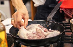Hand placing raw chicken pieces into a hot frying pan on the hob.