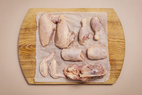 A whole chicken jointed into eight pieces on baking parchment on a wooden board — two breasts, two thighs, two drumsticks, two wings and the stripped carcass — photographed from above on a pale pink background.