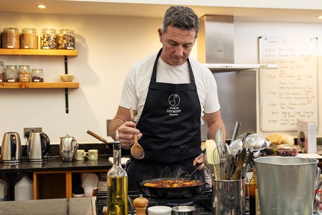Dublin Cookery School student in a DCS apron stirring a cast iron pan on the hob, kitchen shelves and course schedule visible in the background