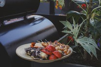 A bamboo plate of freshly grilled food — chicken skewers, charred tomatoes, radicchio and peppers — resting on the side shelf of a large black barrel smoker, surrounded by garden plants in bloom.