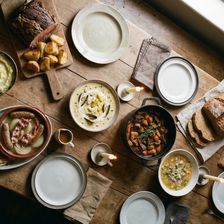 A overhead shot of a rustic farmhouse table laid with a full spread of traditional Irish dishes including brown soda bread, beef and Guinness stew in a cast iron pot, seafood chowder, roast beef with crispy potatoes, mashed potato with butter, Dublin coddle and bacon and cabbage with whiskey caramel sauce, lit by tallow candles and natural window light.