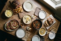 A overhead shot of a rustic farmhouse table laid with a full spread of traditional Irish dishes including brown soda bread, beef and Guinness stew in a cast iron pot, seafood chowder, roast beef with crispy potatoes, mashed potato with butter, Dublin coddle and bacon and cabbage with whiskey caramel sauce, lit by tallow candles and natural window light.