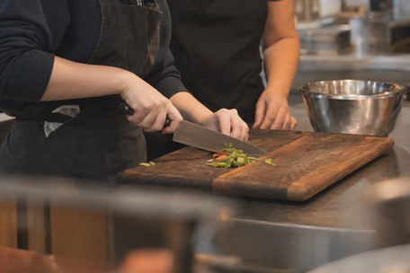 Student practising knife skills on a wooden chopping board at a professional kitchen station, with an instructor's hands visible guiding the technique