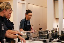 Two students in Dublin Cookery School aprons working with a tutor at the hob during an evening class, one stirring a pan with a wooden spoon, mixing bowls and stockpots along the bench in front of them.