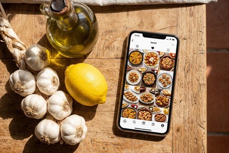 A string of garlic, a jug of olive oil and a lemon on a sunlit wooden table, beside a phone showing a folder of saved Spanish tapas recipes."