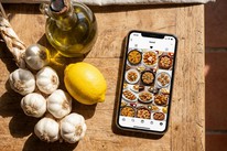 A string of garlic, a jug of olive oil and a lemon on a sunlit wooden table, beside a phone showing a folder of saved Spanish tapas recipes."