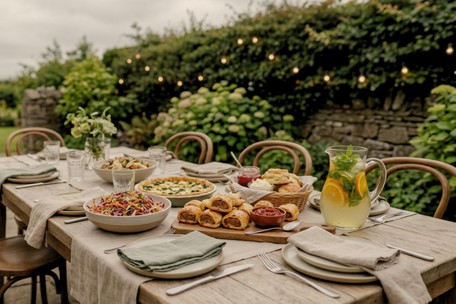A summer garden party spread on an outdoor table at Dublin Cookery School in Blackrock: rainbow slaw, a spinach and feta tart, sausage rolls with tomato relish, and a jug of herb-and-citrus mocktail.