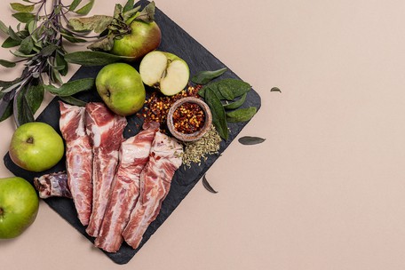 Raw Irish pork ribs on a black slate board with Bramley apples, fresh sage, fennel seeds and chilli flakes, on a soft pink background.