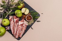 Raw Irish pork ribs on a black slate board with Bramley apples, fresh sage, fennel seeds and chilli flakes, on a soft pink background.