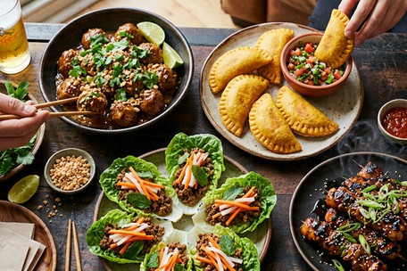 A watch party spread on a wooden table — Thai pork meatballs in a dark bowl, golden empanadas with a salsa dipping bowl, Malaysian-style lettuce cups, and glazed yakitori skewers, with a cold beer in frame and two hands reaching in.