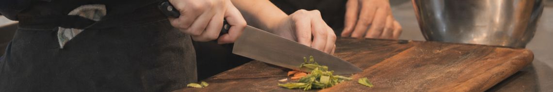 Close-up of hands using a chef's knife on a dark wooden board during a supervised cooking class at Dublin Cookery School