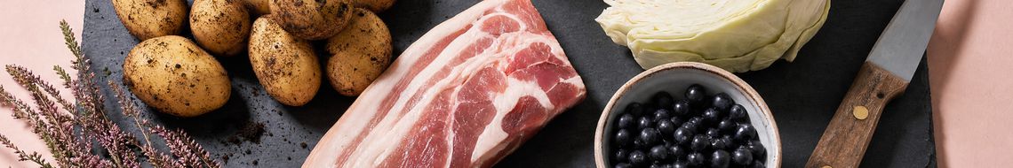 Raw ingredients for the traditional Irish Lúnasa meal arranged on a dark slate board — freshly dug new potatoes with soil still on their skins, a piece of bacon collar, a quartered white cabbage and a small bowl of dark fraughans, with a sprig of heather.