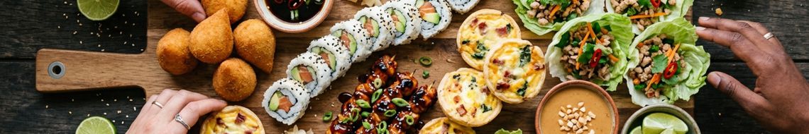 Top-down view of a World Cup finger food spread on a dark wooden board — Brazilian coxinha, sushi rolls, yakitori skewers, mini quiches, lettuce cups with chilli, and peanut dipping sauce. Hands reaching in from three sides.
