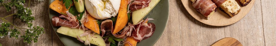 A professional overhead shot of an abundant spread of budget-friendly party recipes on a rustic wooden table. The selection features inexpensive party food ideas including mini croque monsieurs, Parma ham and rocket rollups, and sautéed prawns with cherry tomatoes. These dishes represent cheap party food to feed a crowd for Six Nations Super Saturday in Ireland.