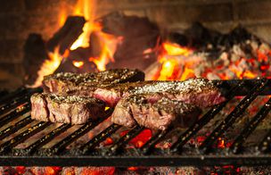 A shared barbecue feast on a wooden board — grilled meats, charred vegetables and prosciutto, with bowls of cherry tomatoes and dressings, as hands reach in to serve — Dublin Cookery School's Summer BBQ Feast.