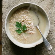 Overhead shot of a bowl of homemade chicken and barley broth in a worn ceramic bowl, shredded chicken and pearl barley visible beneath the surface, garnished with a single sprig of flat leaf parsley, with a silver spoon resting in the bowl on a rough stone surface.