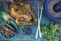 Chargrilled whole chicken with golden, crispy skin served alongside grilled baby aubergines, fresh lemongrass, kaffir lime leaves, coriander, and a mortar of nam jim sauce on a rustic green wooden table. A colourful outdoor summer cooking scene.
