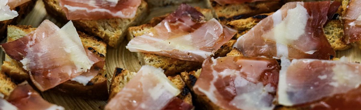 Pieces of toasted bread topped with folds of cured ham and shavings of cheese, on a wooden board