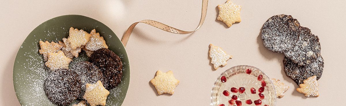 Star and Christmas tree biscuits and chocolate biscuits dusted with icing sugar, beside a glass of pomegranate seeds