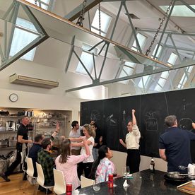 A group of adults in a bright, high-ceilinged kitchen with steel roof trusses and skylights, gathered around a large blackboard covered in chalk drawings during what appears to be a team-based cooking challenge.