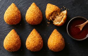 Six golden Brazilian Coxinha on a dark slate board, one broken open to show the shredded chicken filling, with a small bowl of red dipping sauce and flour dusting on the surface. Dublin Cookery School recipe by tutor Rafa.