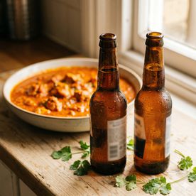 Two cold beer bottles with condensation beside a bowl of summer curry on a wooden counter — Summer Curries and Beer evening class at Dublin Cookery School