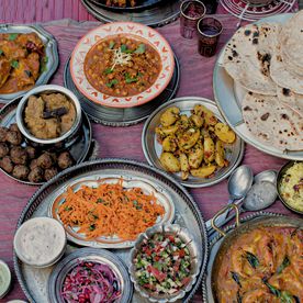 An overhead shot of a generously laid Indian-inspired feast on a pink cloth — chickpea curry, prawn masala, spiced potatoes, meatballs, shredded carrot salad, raita, flatbreads and various chutneys served in traditional silver and ceramic dishes.