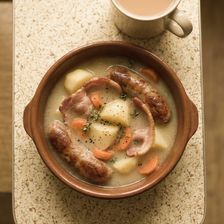 Overhead shot of a traditional Dublin coddle served in a terracotta dish — pork sausages, bacon rashers, soft potatoes and sliced carrots in a pale onion broth, garnished with fresh thyme, with a mug of tea visible at the top of the frame.