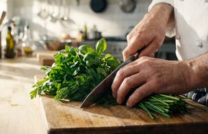 Hands chopping fresh herbs on a wooden board with a chef’s knife.