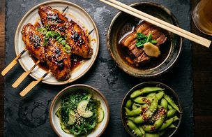 Four izakaya-style small plates on a dark slate board — chicken wing yakitori with sticky soy glaze, sesame and spring onion on skewers; umami pork belly braise in a dark ceramic bowl with chopsticks; seaweed and cucumber salad with sesame; edamame with chilli flakes. A glass of beer at the edge of the frame. Dishes from the Japanese Fusion Night at Dublin Cookery School, Blackrock.