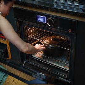 A person placing a black cast-iron casserole dish into a modern oven in a home kitchen, preparing for slow cooking. The scene shows wooden floors, stainless-steel knobs, and natural light, suggesting a warm, lived-in space ready for winter cooking.