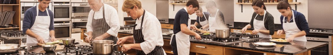 A group of students and instructors cooking together in a bright, modern teaching kitchen with skylights and open shelving. Each person is focused on preparing food at large wooden workstations with stainless-steel stoves, reflecting a hands-on cookery class environment.