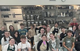A group of students in black aprons collaborating over a steaming pot during a hands-on cookery class, listening to instruction in a busy kitchen environment.