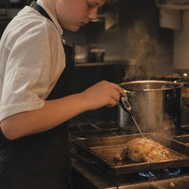 Teenager using a digital probe thermometer to check the internal temperature of roast chicken on a gas hob, with steam rising in a professional kitchen