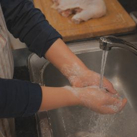 Teenager washing hands with soap under running water at a stainless steel kitchen sink, with raw chicken visible on a chopping board in the background