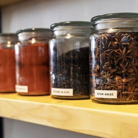 Glass jars neatly arranged on a wooden kitchen shelf, filled with winter spices and ingredients including star anise, black sesame seeds, and paprika. Each jar is clearly labelled, reflecting an organised and practical pantry setup.