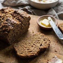 A sliced loaf of Irish brown soda bread on a wooden chopping board with sea salt crystals on the crust, a cut slice in the foreground showing the dense wholemeal crumb, a small dish of butter with a knife resting beside it and a bowl of seafood chowder softly blurred in the background.
